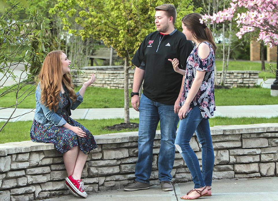 Three people, two standing and one sitting on a stone ledge, engaging in conversation outdoors near blooming trees and greenery.